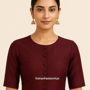 May include: A maroon blouse with short sleeves and a round neckline. The blouse has a button-down front and the text "TrishanFashionHub" printed on the front. The model is wearing gold earrings.