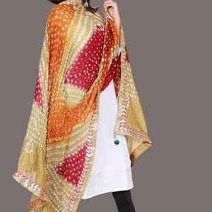 May include: A colorful, patterned scarf in shades of gold, orange, and red. The scarf has a detailed design with small white dots and a decorative border. The fabric drapes over the shoulders, creating a flowing effect. The scarf is a vibrant accessory.