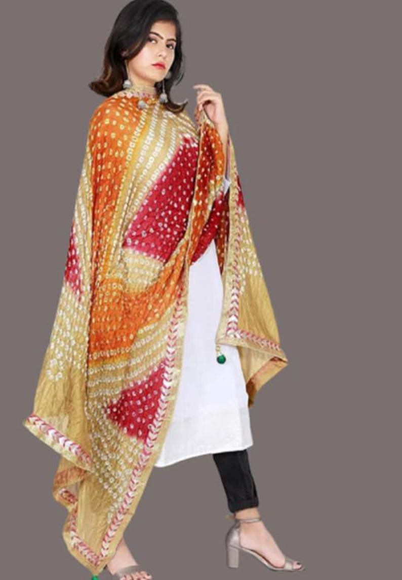 May include: A colorful, patterned scarf in shades of gold, orange, and red. The scarf has a detailed design with small white dots and a decorative border. The fabric drapes over the shoulders, creating a flowing effect. The scarf is a vibrant accessory.