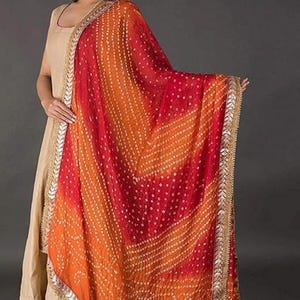 May include: A vibrant, patterned scarf in shades of red and orange, with a decorative border. The scarf features a detailed design and is likely made of a lightweight fabric. The scarf is a fashion accessory, suitable for adding a pop of color to an outfit.