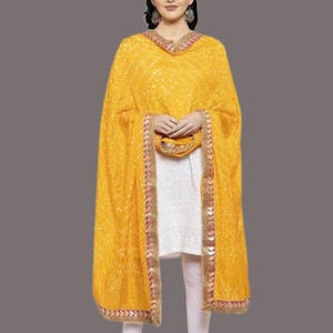 May include: A woman wearing a yellow dupatta with a white outfit. The dupatta has a detailed pattern and a decorative border with red and gold accents. The outfit includes a white tunic and white leggings.