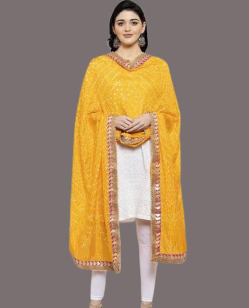 May include: A woman wearing a yellow dupatta with a white outfit. The dupatta has a detailed pattern and a decorative border with red and gold accents. The outfit includes a white tunic and white leggings.