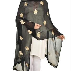 May include: A black sheer scarf with gold embroidery and black tassels. The scarf is draped over a person's shoulders and is worn with a white top and white pants.