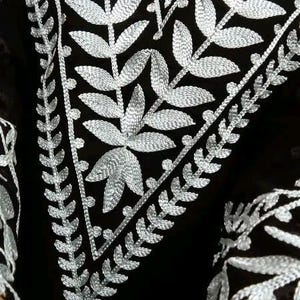 May include: Close-up of a black garment with intricate silver embroidery. The embroidery features a leaf and vine pattern, creating a detailed and elegant design. The garment's dark color contrasts with the bright silver thread.
