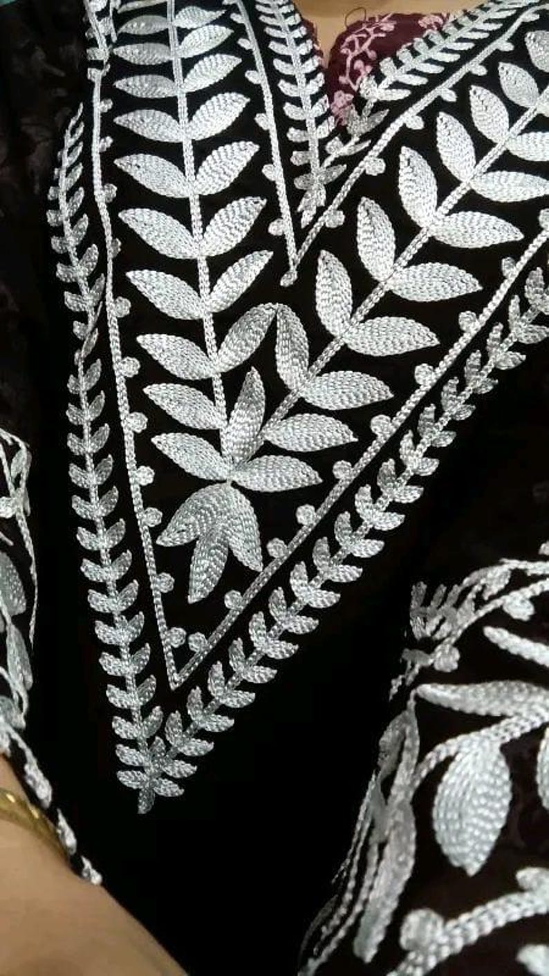May include: Close-up of a black garment with intricate silver embroidery. The embroidery features a leaf and vine pattern, creating a detailed and elegant design. The garment's dark color contrasts with the bright silver thread.