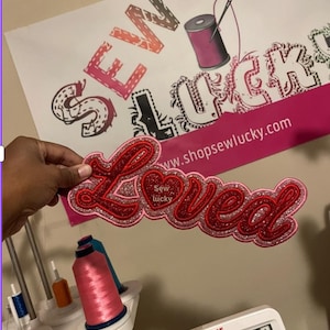 May include: A red and glittery "Loved" applique with a heart and the text "Sew lucky". The applique is held in front of a poster with the text "Sew Lucky" and a website address. Sewing supplies are visible in the background.