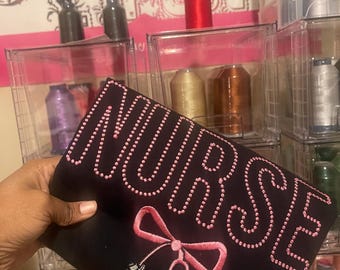 Pode incluir: Uma bolsa de tecido preta com a palavra "NURSE" bordada em rosa com strass rosa delineando as letras. Uma fita rosa com um estetoscópio está bordada abaixo da palavra.
