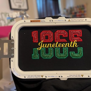 May include: Black fabric embroidered with the words "10CC Juneteenth LUUJ" in red, yellow, and green glitter thread. The design is held in a white embroidery hoop.