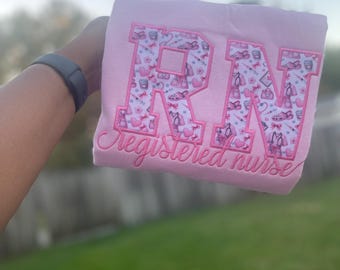 May include: A light pink sweatshirt with the embroidered letters "RN" in a pattern of medical supplies and hearts. The words "Registered nurse" are embroidered below the letters. The sweatshirt is held up against a green background.