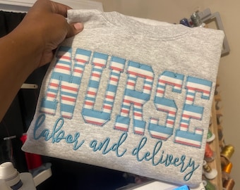May include: A gray sweatshirt with the word "NURSE" embroidered in blue and pink stripes. The word "labor and delivery" is embroidered below in blue.