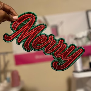 May include: A Christmas-themed patch with the word "Merry" in a cursive font. The word is red with glitter, outlined in red, and set against a green background. The patch is likely for clothing or accessories.
