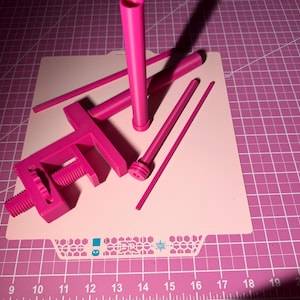 May include: A bright pink 3D printed assembly consisting of a C-clamp with a threaded screw mechanism, a tall vertical cylinder, and three slender rods of varying lengths. The components are arranged together on a flat, light-colored surface.