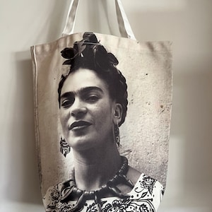 Può includere: Una borsa tote in tela bianca con un'immagine in bianco e nero di Frida Kahlo stampata sul davanti. L'immagine mostra il viso e le spalle di Frida Kahlo, con la sua corona di fiori e i suoi gioielli caratteristici.