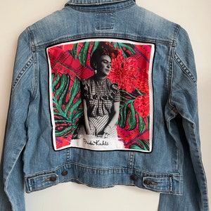 May include: A blue denim jacket with a colourful patch on the back featuring a portrait of Frida Kahlo with the text "Frida Kahlo" underneath.