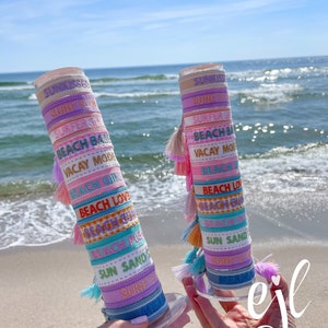 May include: Two stacks of colorful bracelets with tassels, each stack has a different beach-themed phrase on each bracelet. The phrases include "Sunkissed", "Surfer Girl", "Beach Babe", "Vacay Mode", "Beach Love", "Beach Bum", "Beach Pile", "Sun Sand", and "Surf's Up".