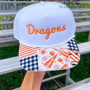 May include: A white trucker hat with a mesh back and a white rope detail across the front. The word "Dragons" is embroidered in orange script. The brim features a patchwork pattern of orange stripes, black and white gingham, and football icons.