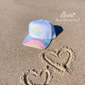 May include: White trucker hat with a rainbow-colored brim and the text "BE THE LIGHT Matthew 5:14". The hat is on a sandy beach with two hearts drawn in the sand. The brand name "elleete" is also visible.