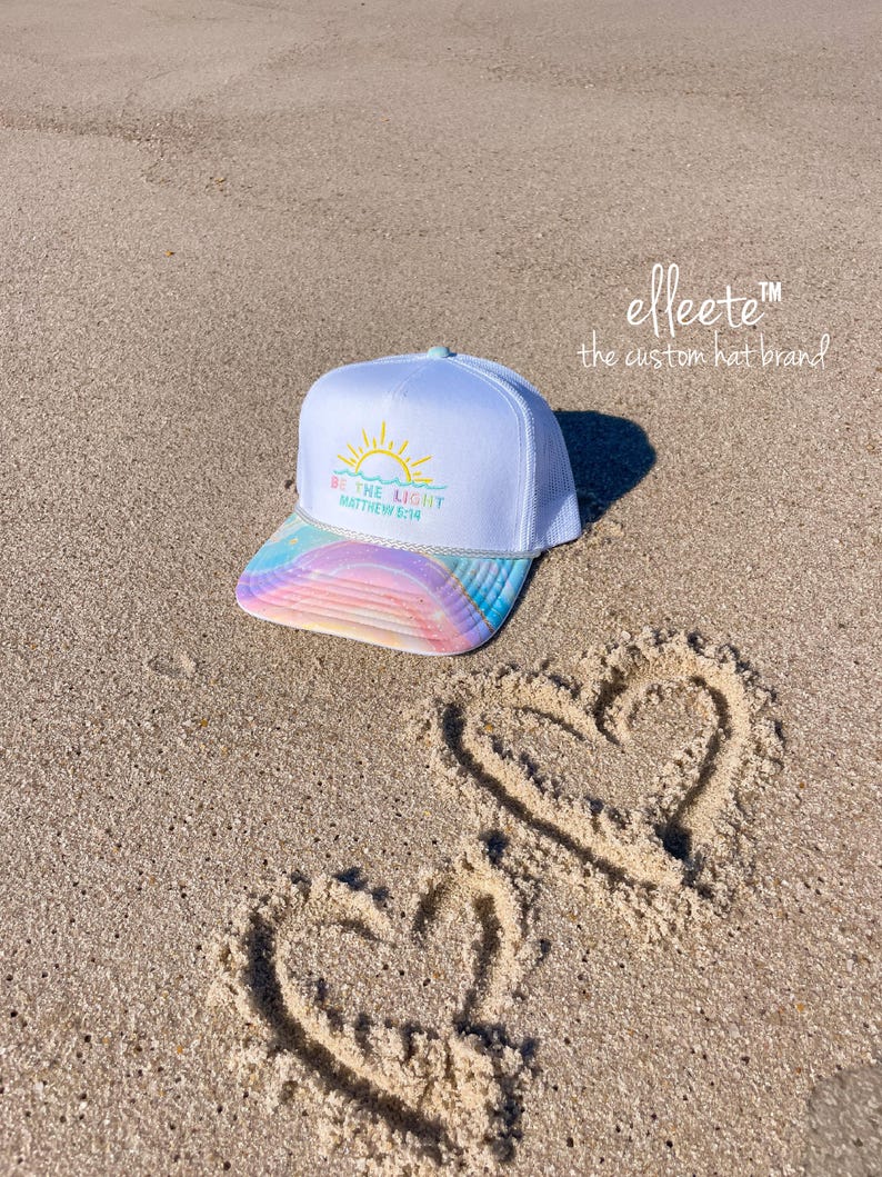 May include: White trucker hat with a rainbow-colored brim and the text "BE THE LIGHT Matthew 5:14". The hat is on a sandy beach with two hearts drawn in the sand. The brand name "elleete" is also visible.