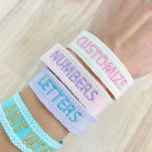 以下が含まれることがあります： 刺繍されたテキストが施された4本のカラフルな織りブレスレット。ブレスレットは人の手首に重ねられています。ブレスレットのテキストは「Any Word」、「Letters」、「Numbers」、「Customize」です。