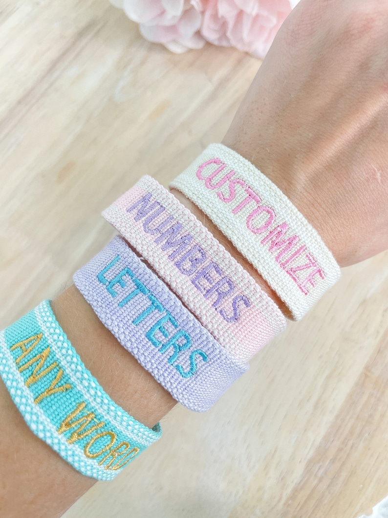 以下が含まれることがあります： 刺繍されたテキストが施された4本のカラフルな織りブレスレット。ブレスレットは人の手首に重ねられています。ブレスレットのテキストは「Any Word」、「Letters」、「Numbers」、「Customize」です。