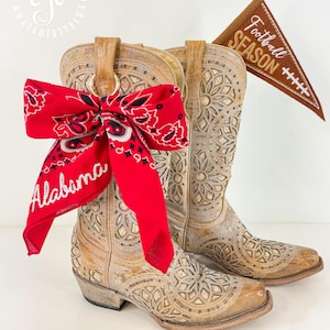 May include: A pair of tan leather cowboy boots with intricate floral cutouts. A red bandana with "Alabama" text is tied to one boot. A brown pennant with "Football Season" text is tucked into the other boot.