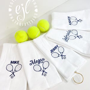 May include: Six white towels with embroidered tennis rackets and a tennis ball. Each towel has a different name embroidered on it: Mike, Megan, Jerry, Gabriela, Andrew. Three yellow tennis balls are in the background.