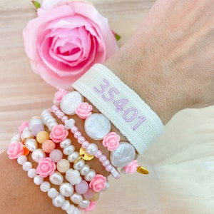 May include: A stack of bracelets with pink flowers, white pearls, and a white fabric wristband with the text "35401" in purple.