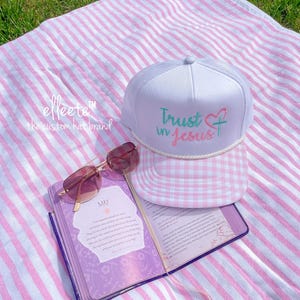 May include: A white trucker hat with a pink and white gingham brim and the text "Trust in Jesus" in teal and pink. The hat is on a pink and white striped blanket with a pair of sunglasses and a purple journal.
