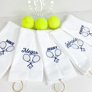 May include: Five white towels with gold loops, each embroidered with a name and two crossed tennis rackets. The names are Mike, Megan, Jerry, Gabriela, and Andrew. Three yellow tennis balls are in the background.