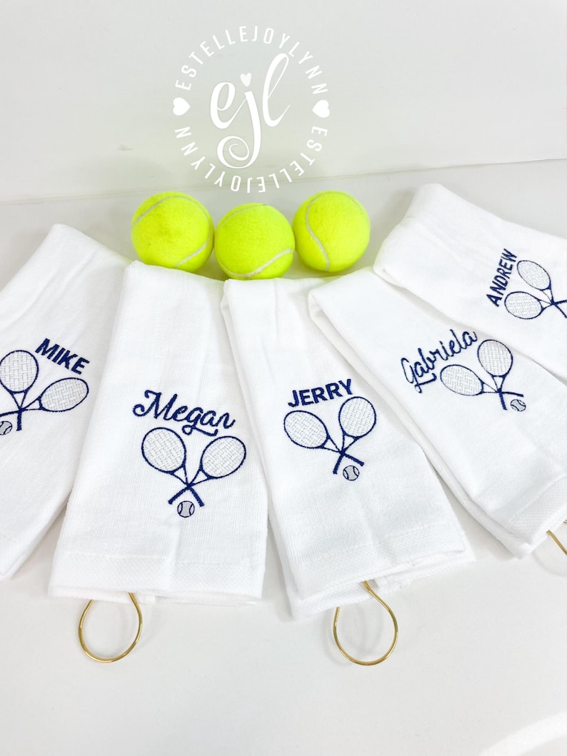 May include: Five white towels with gold loops, each embroidered with a name and two crossed tennis rackets. The names are Mike, Megan, Jerry, Gabriela, and Andrew. Three yellow tennis balls are in the background.
