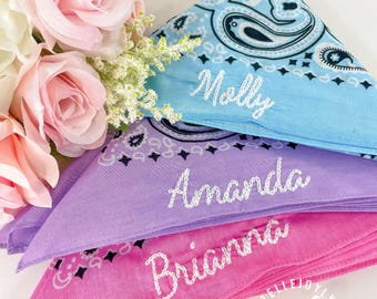 May include: Three personalized bandanas in blue, purple, and pink, each embroidered with a name in white script: Molly, Amanda, and Brianna. The bandanas feature a paisley design and are folded into triangles. Pink roses and white flowers are in the background.