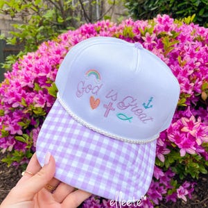 May include: White trucker hat with a lavender gingham brim. The hat features the embroidered text "God is Great" with a rainbow, heart, cross, fish, and anchor. The brand name "effeete" is printed on the brim.