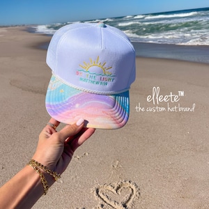 May include: White trucker hat with a colorful, pastel-colored bill and the text "BE THE LIGHT MATTHEW 5:16" embroidered on the front. The hat features a sun design and is held up on a sunny beach.