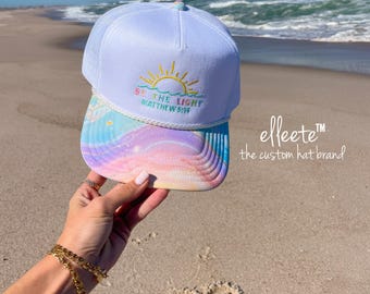 May include: White trucker hat with a colorful, pastel-colored bill and the text "BE THE LIGHT MATTHEW 5:16" embroidered on the front. The hat features a sun design and is held up on a sunny beach.