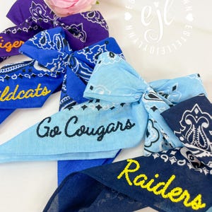 Puede incluir: Una colecci&oacute;n de cuatro lazos para el pelo de tela con estampado de bandana en tonos morado, azul real, azul claro y azul marino. Los lazos presentan texto bordado que incluye Tiger, Wildcats, Go Cougars y Raiders en amarillo o negro.