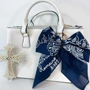 May include: A white handbag with a top handle and gold-colored hardware. A navy blue bandana with a white paisley design and the text "Romans 8:28" is tied to the handle. A white cross with a rhinestone is on the left.