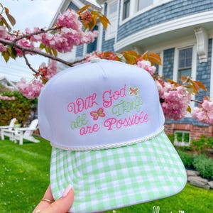 May include: White trucker hat with a green and white gingham brim. The hat features the embroidered text "With God all things are Possible" in pink and green, with butterfly accents. The hat is from the brand "elletee".