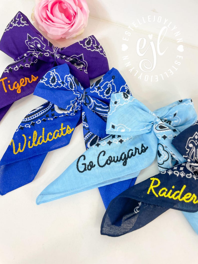 Puede incluir: Cuatro lazos de tela hechos de material de bandana en morado, azul real, azul claro y azul marino. Cada lazo presenta un patr&oacute;n de cachemira blanco y texto bordado: 'Tigers' en amarillo, 'Wildcats' en amarillo, 'Go Cougars' en negro y 'Raiders' en amarillo.