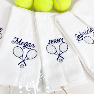 May include: Four white towels with embroidered tennis rackets and names: Megan, Jerry, Gabriela. Each towel has a gold ring attached. Three yellow tennis balls are in the background.