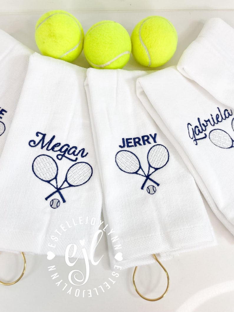 May include: Four white towels with embroidered tennis rackets and names: Megan, Jerry, Gabriela. Each towel has a gold ring attached. Three yellow tennis balls are in the background.