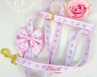 May include: A pink and white gingham dog leash set with a matching bow tie and a pink rose. The leash and bow tie feature a floral pattern and the name "Dixie" embroidered in pink. Gold-colored hardware.