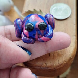 Pode incluir: Uma pequena figura de sapo feita à mão de argila polimérica. O sapo é pintado em tons de rosa, azul e roxo com olhos pretos.