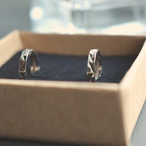 May include: Two silver hoop earrings with a geometric design, displayed in a brown box with a blue velvet lining.