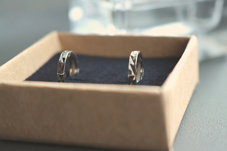 May include: Two silver hoop earrings with a geometric design, displayed in a brown box with a blue velvet lining.