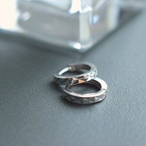 May include: A pair of silver hoop earrings with a geometric pattern. The earrings are on a gray surface.