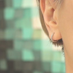 May include: A close-up of a person's ear with a small silver hoop earring. The earring is a simple, thin hoop with a slightly textured surface.