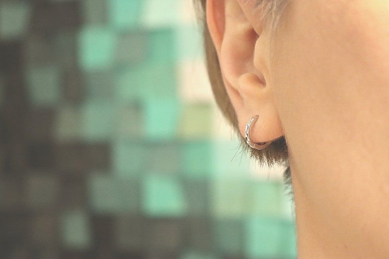 May include: A close-up of a person's ear with a small silver hoop earring. The earring is a simple, thin hoop with a slightly textured surface.