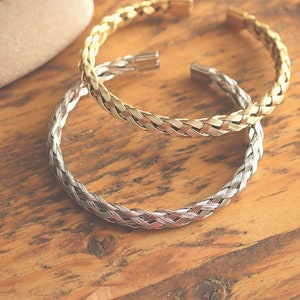 May include: Two braided metal bracelets, one gold and one silver, on a wooden surface.