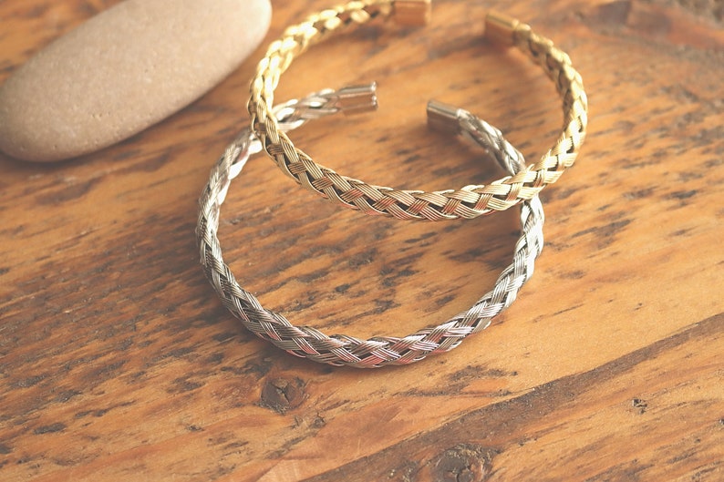 May include: Two braided metal bracelets, one gold and one silver, on a wooden surface.