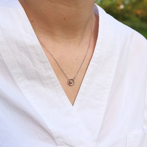 May include: A silver necklace with a small pendant featuring a sphere within a circle. The necklace is worn over a white shirt with a V-neck. The background is blurred, suggesting an outdoor setting.